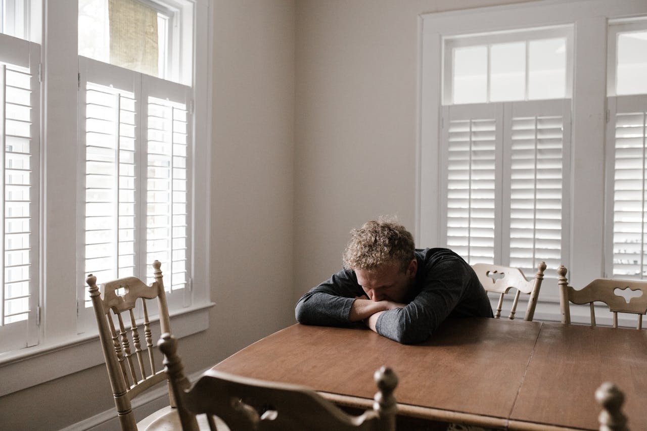 An adult man with emotions of sadness and anxiety sitting alone at a wooden table near windows.